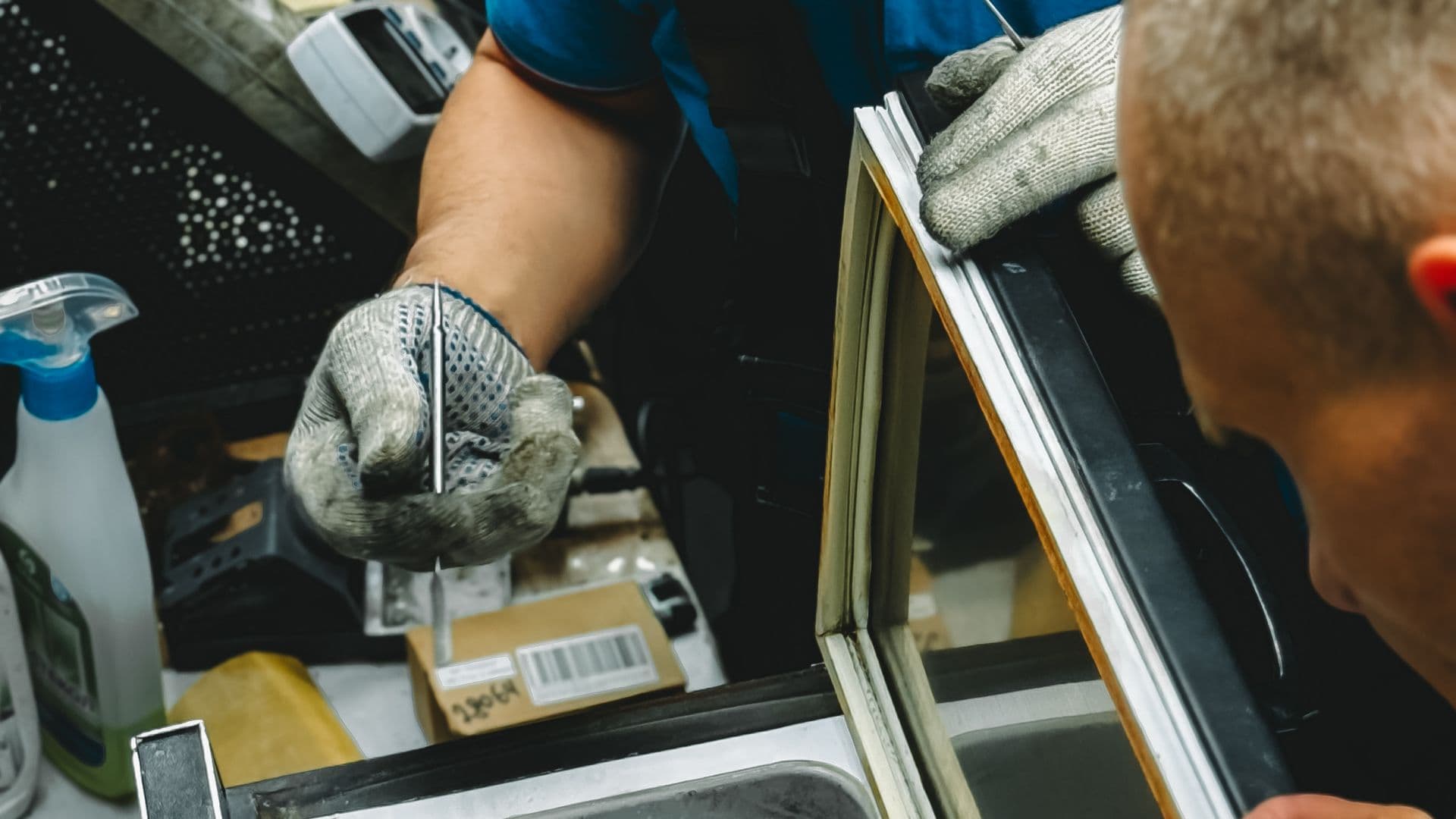 An appliance repair technician kneeling at an open refrigerator with a tool bag, diagnosing in a home kitchen