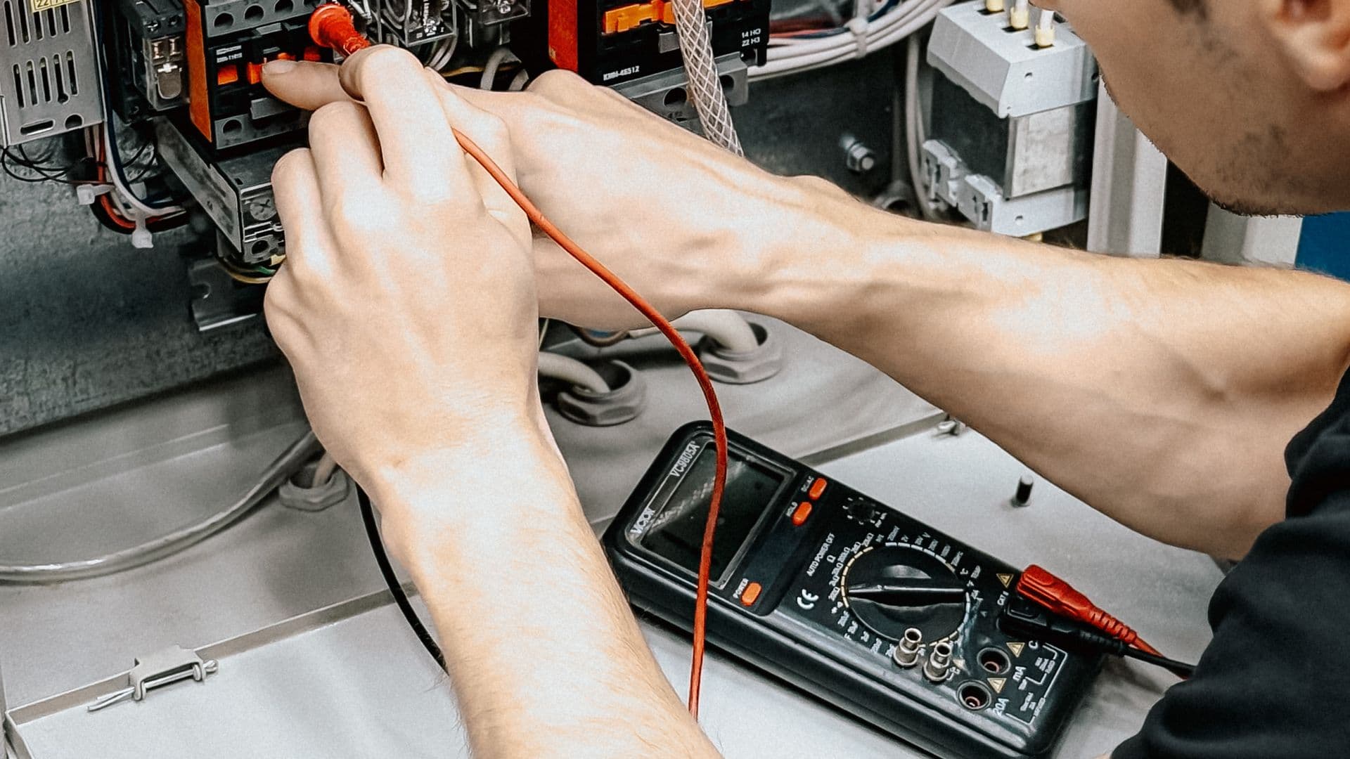 An electrician in a hard hat working inside an open breaker panel with a multimeter, focused, dim utility-room light