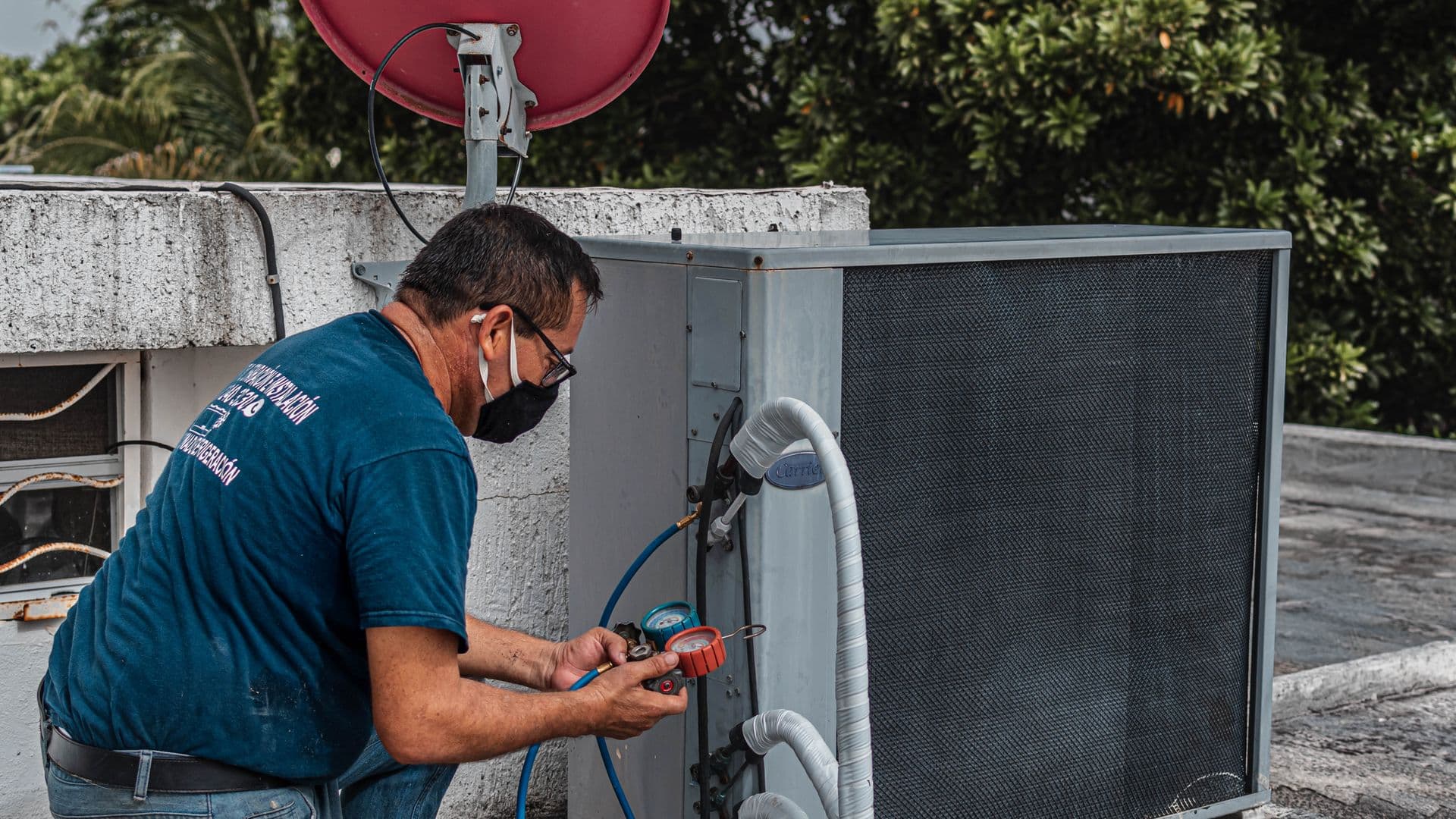 A mature HVAC technician in a face mask kneeling on a rooftop AC unit with refrigerant gauges in hand, residential rooftop