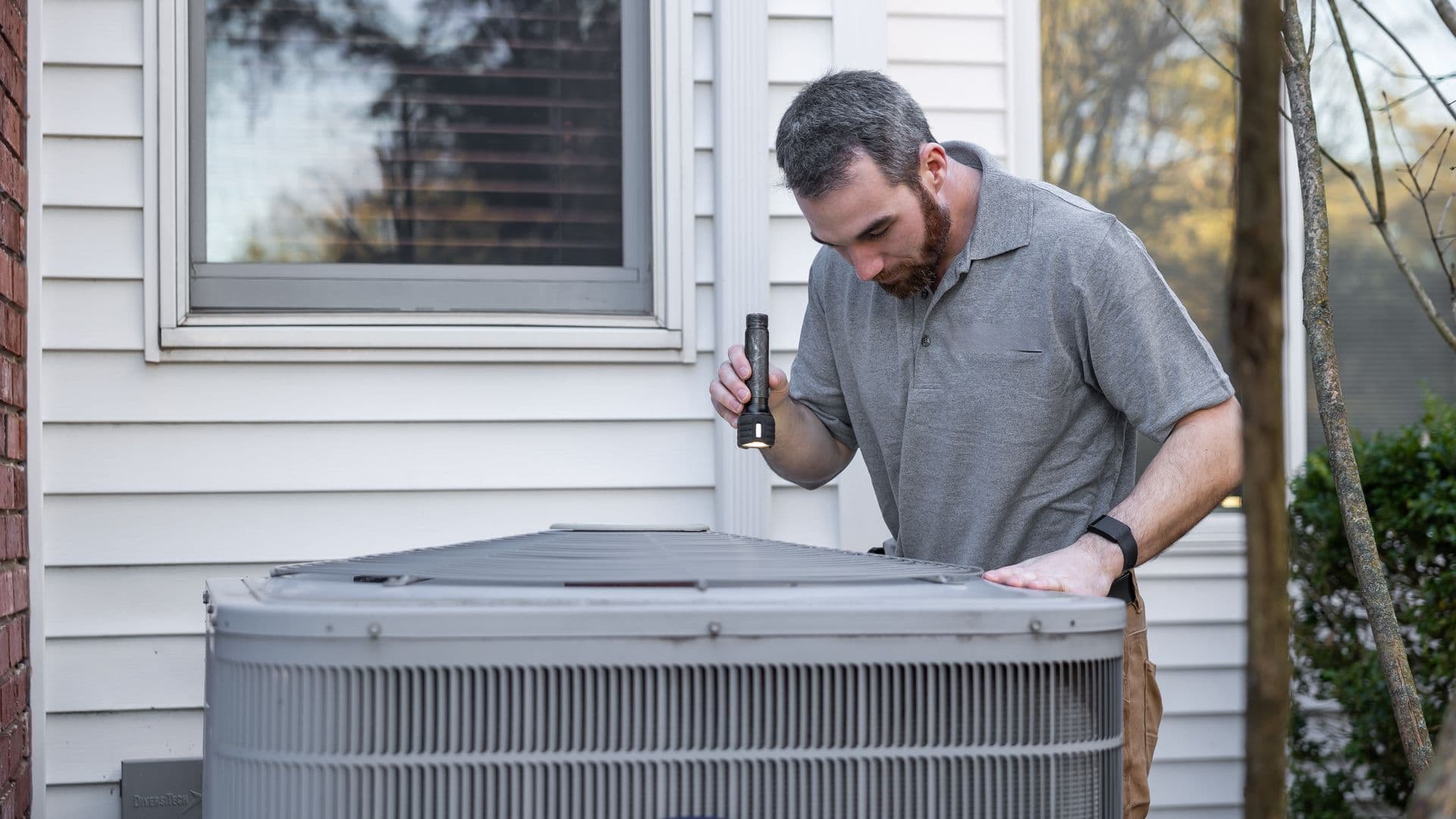 A mature HVAC technician in a gray polo and work belt at a residential outdoor AC unit, leaning over with a flashlight to inspect the coils, suburban siding and a window behind him