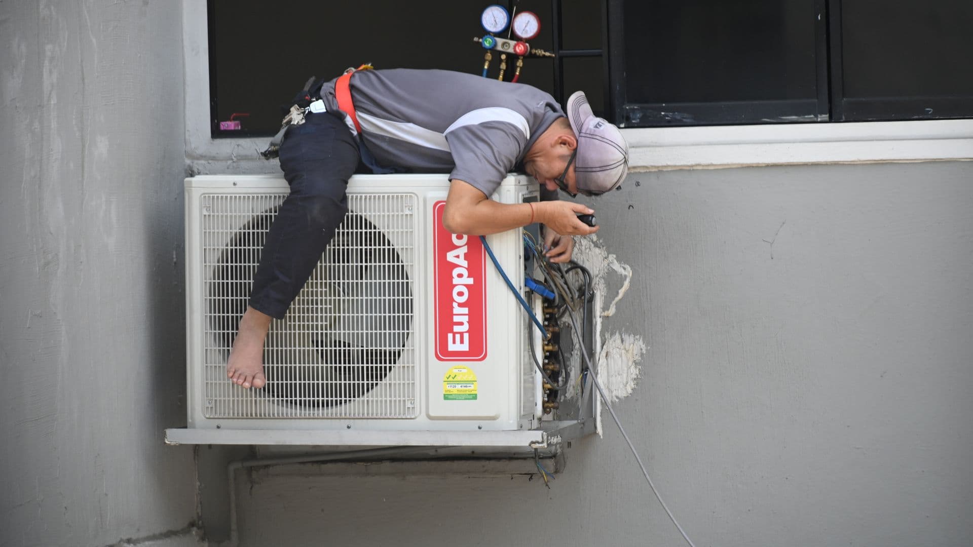 An HVAC technician in a gray uniform leaning across a wall-mounted condenser unit mid-repair, working through peak season