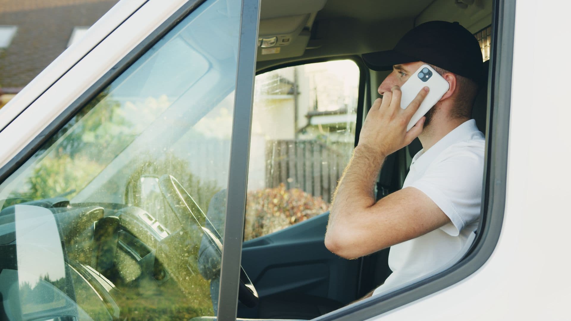 A young tradesman in a white polo and black cap sitting in the cab of a white work van, smartphone held to his ear, summer daylight outside the windshield