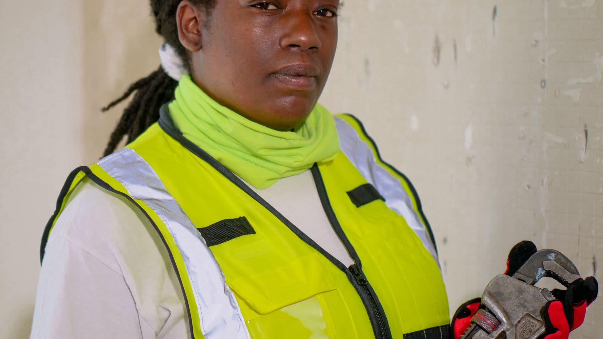 A plumber in a hi-vis safety vest holding a heavy pipe wrench, looking directly at the camera in a partly-built room