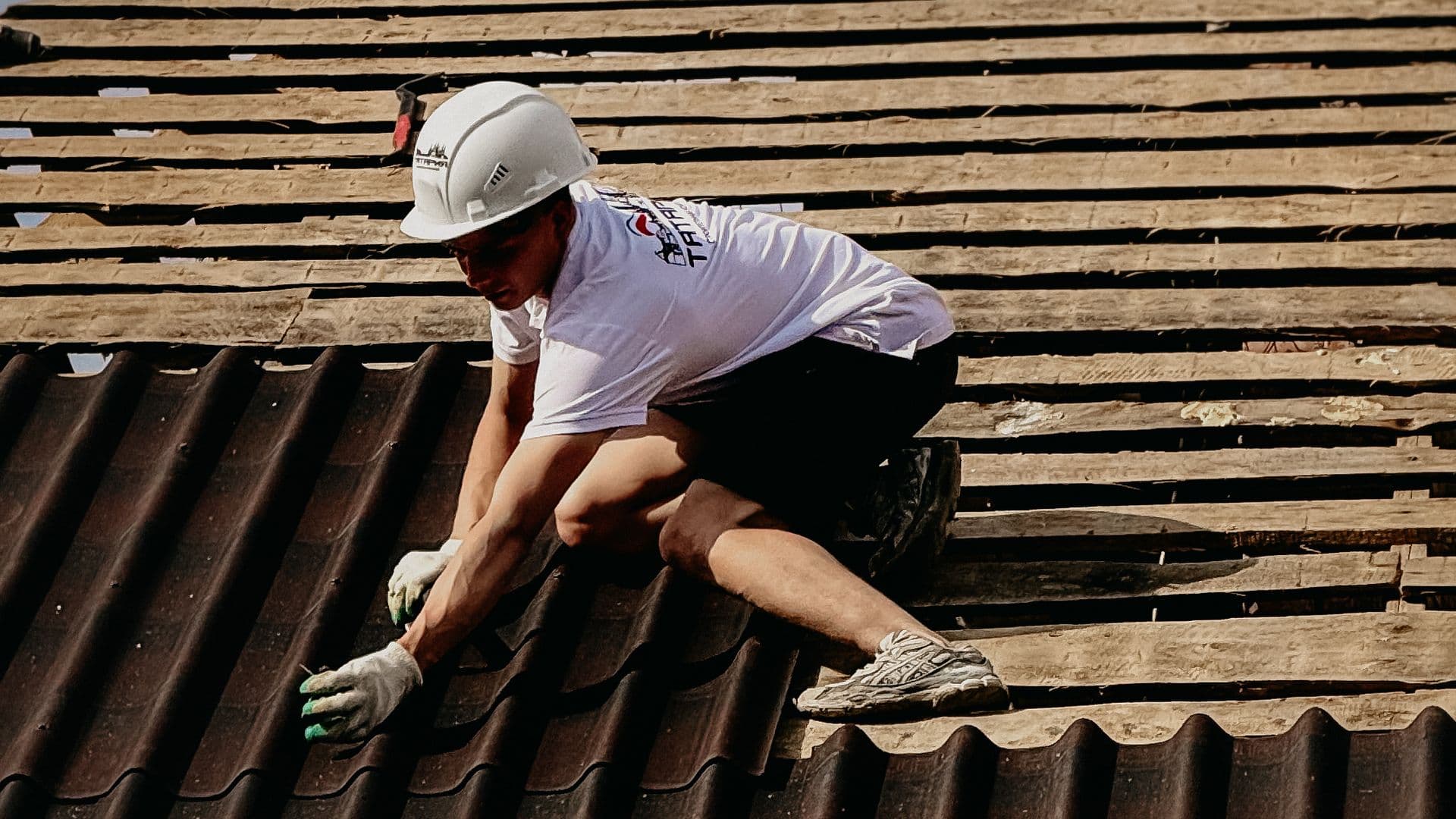 A roofer in a safety harness inspecting hail-damaged shingles on a residential roof under a grey post-storm sky
